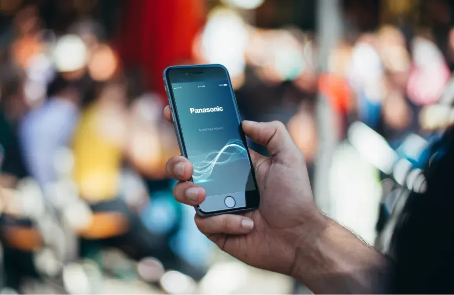 A person holding a phone with the Panasonic Battery App open against a blurred city background.